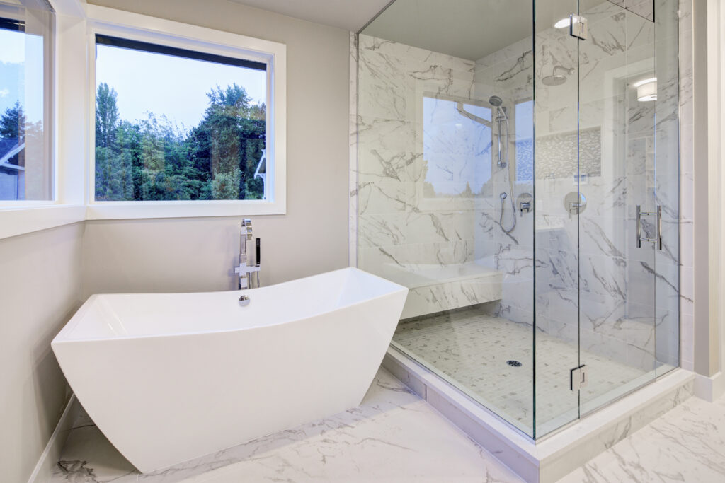 Sleek bathroom features freestanding bathtub atop marble floor placed in front of glass shower accented with rain shower head and gray and white marble surround.